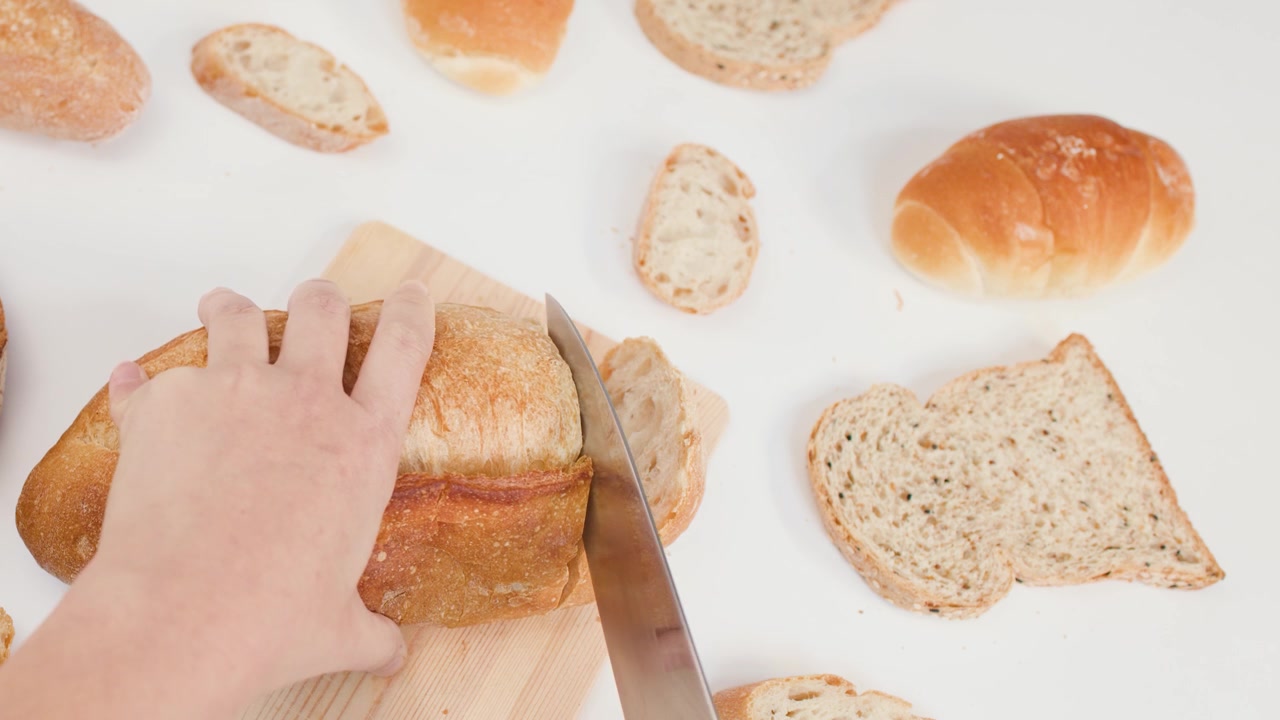 Overhead view of hands slicing organic sourdough loaf on wooden board against white background.