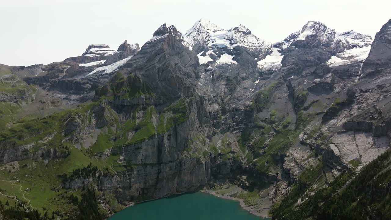 Aerial shot of Oeschinen Lake in Switzerland, tilting upward to reveal the towering cliffs, green slopes, and snow-capped peaks of the Bernese Alps