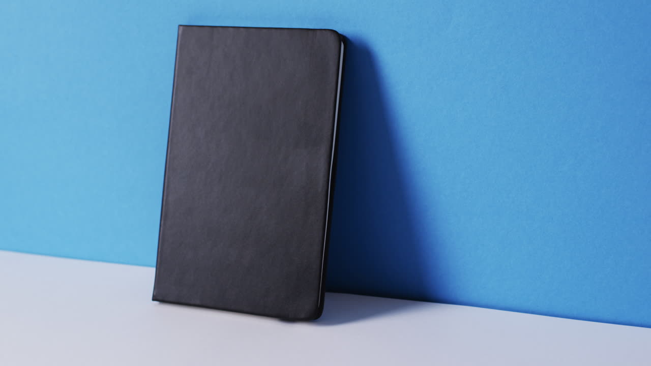 Close up of closed black book leaning on wall with copy space on blue background in slow motion