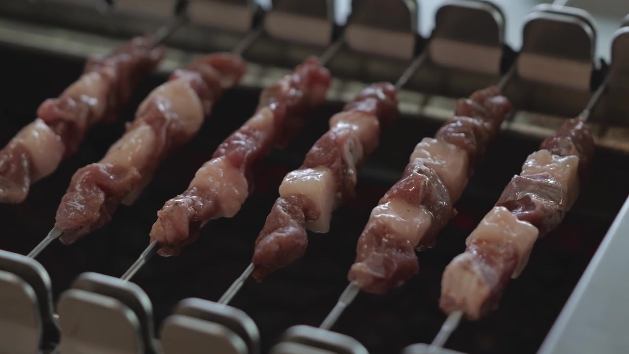 Primo piano di spiedini per barbecue che ruotano lentamente su carboni ardenti su una griglia automatica, catturando cibo che sfrigola, fumo e una texture grigliata appetitosa.