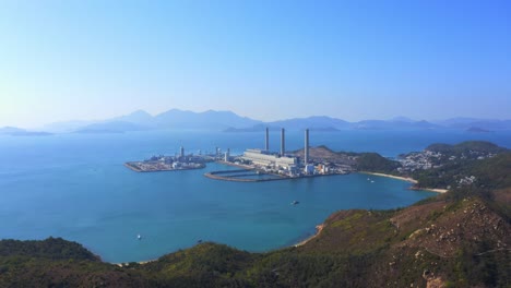 Drone-shot,-traveling-backward,-over-a-big-green-mountain-with-big-coastal-coal-fired-power-plant-on-an-island-next-to-a-village-with-a-beach-during-a-beautiful-sunny-day