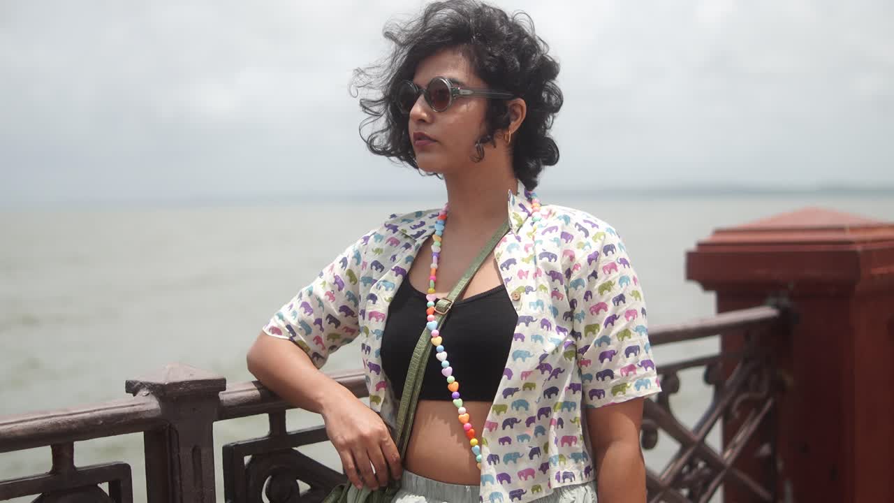 Young Indian woman standing by a waterfront railing, curly hair blowing in the sea breeze as she gazes away, relaxed and carefree on a sunny seaside promenade
