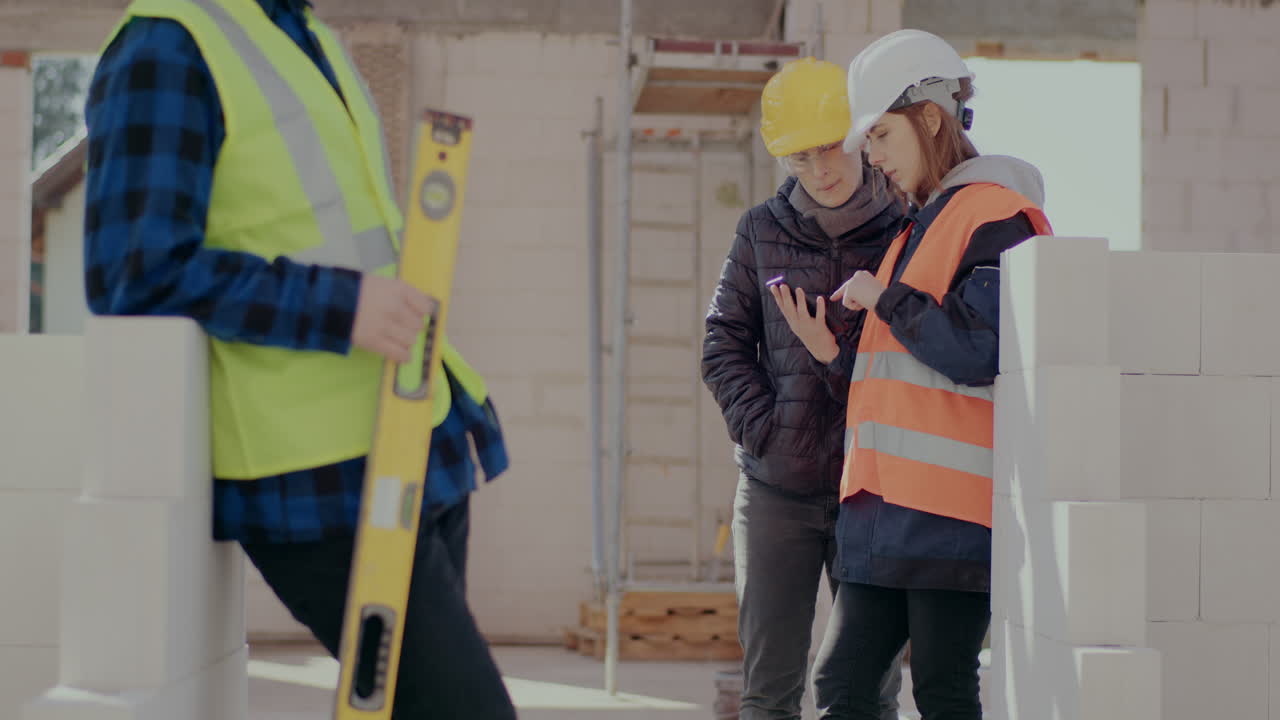 Midsection of male worker holding level standing near female coworker discussing on smartphone with engineer at construction site