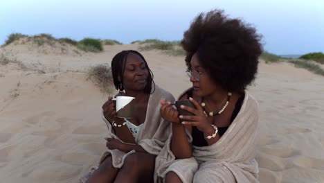 Cheerful-black-girlfriends-drinking-coffee-on-beach