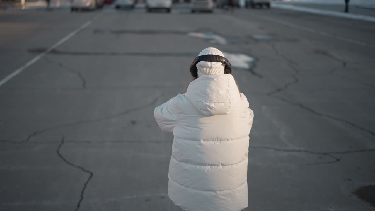 Back view of young lady in white winter coat and beanie moving rhythmically on wide urban sidewalk during sunny winter day, enjoying peaceful moment with headphones under clear blue sky