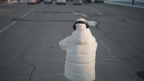 back view of young lady in white winter coat and beanie moving rhythmically on wide urban sidewalk during sunny winter day, enjoying peaceful moment with headphones under clear blue sky