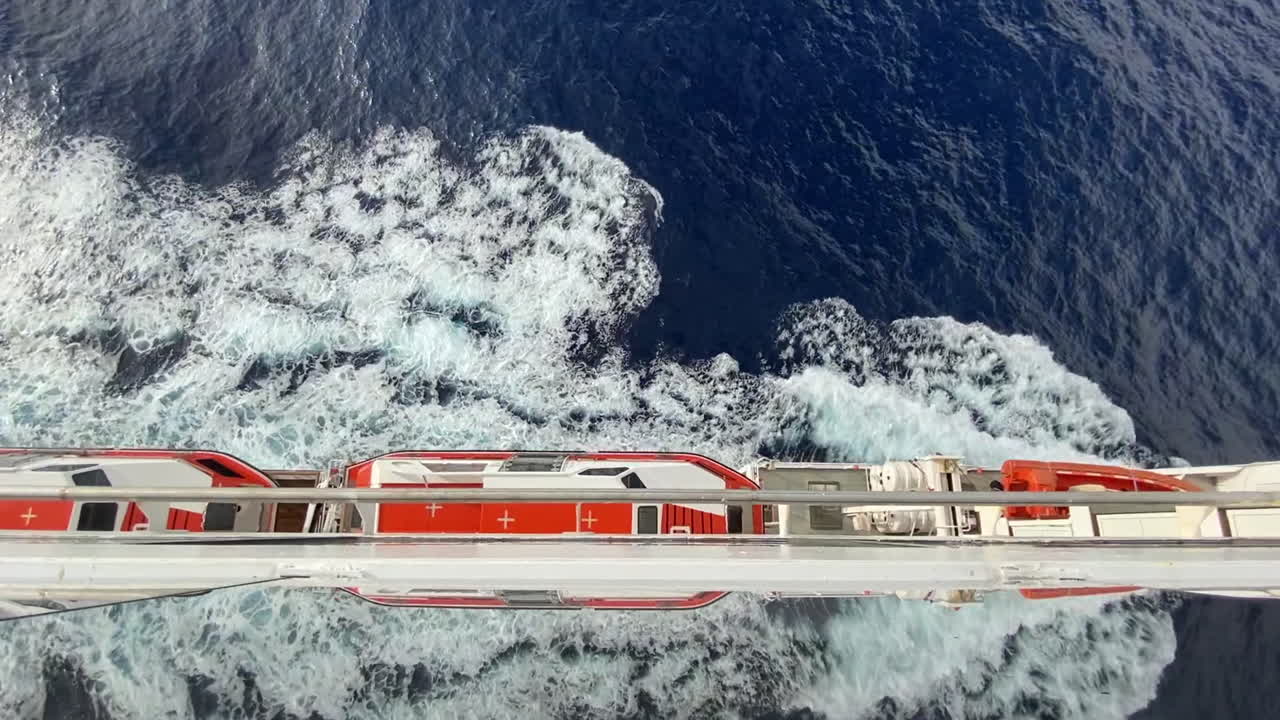 orange lifeboats on Portside of a cruise ship on a voyage with waves rooling back to the sea with a mirror view. Atop wide steady shot