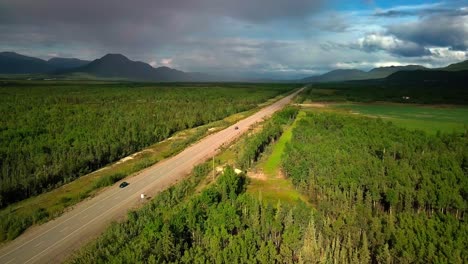 vuelo panorámico de verano sobre la estrecha y estrecha carretera de alaska en el valle de ibex por árboles verdes siempre verdes en el bosque hacia la cordillera, nubes lluviosas y cielo azul, enfoque aéreo superior