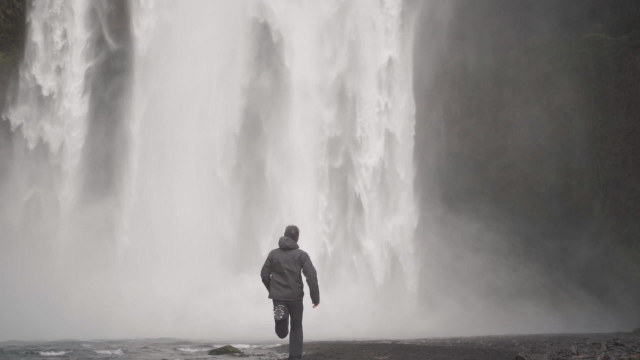 Una toma dinámica de un joven corriendo con entusiasmo hacia la majestuosa cascada Skogafoss en Islandia