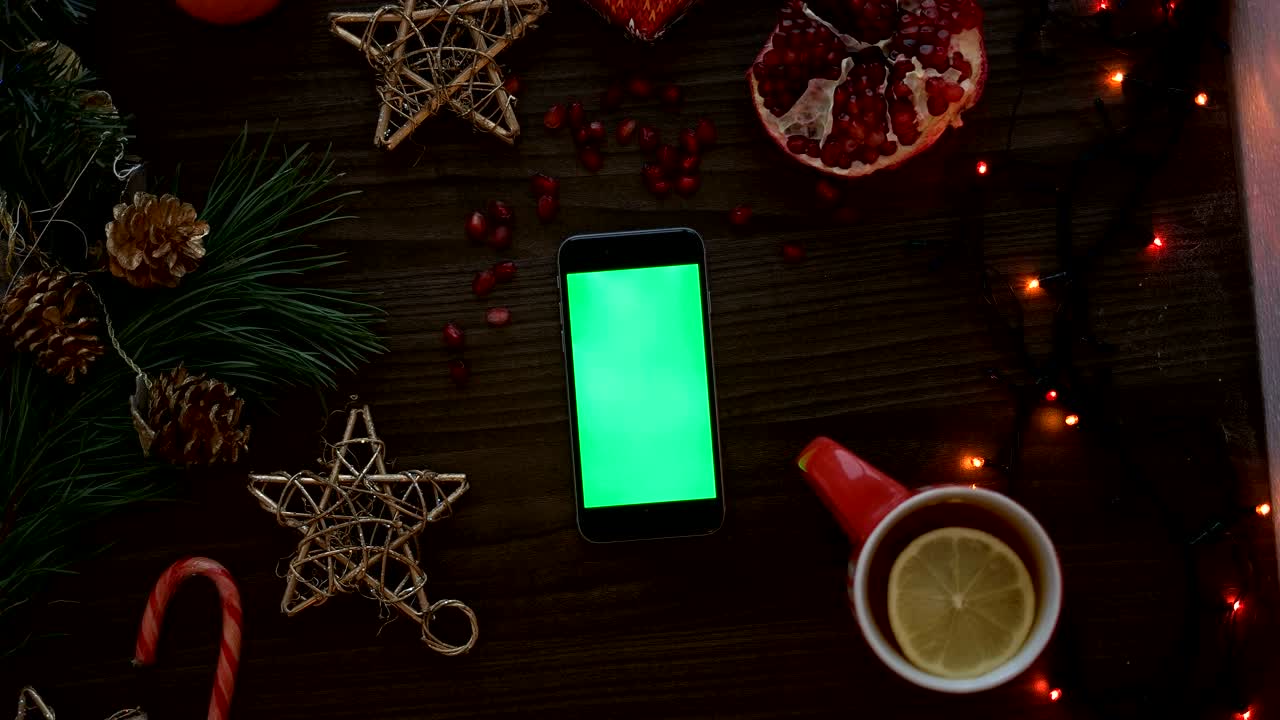 녹색 화면으로 스마트 폰을 사용하여 손으로 위쪽을 보십시오. 터치 스크린에서 손가락 스크롤 페이지. 나무 테이블 배경에서 크리스마스 세부 사항. 크로마 키