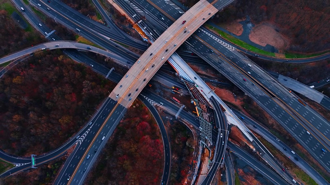 Crossing overpass above a wide freeway with construction, New Jersey. Top-down of an arching flyover spanning multilane traffic next to a bridge building site and red autumn brush