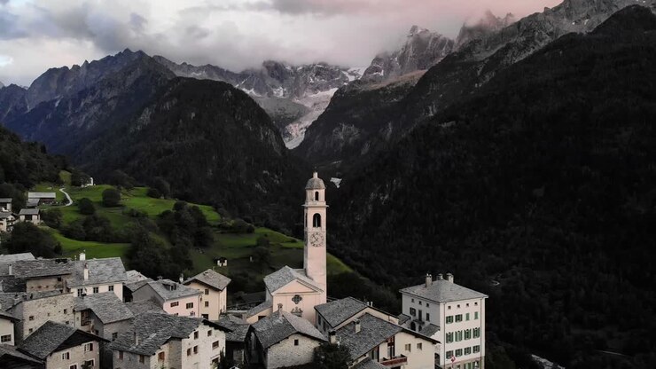Aerial flyover over the rooftops of the historical village of Soglio in the Bregaglia region of Grissons, Switzerland with a view of the old church and mountain peaks of Engadin