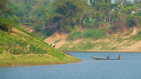 a farmer in bangladesh tends to his crops on the riverbank as a wooden boat with fishermen slowly drifts by