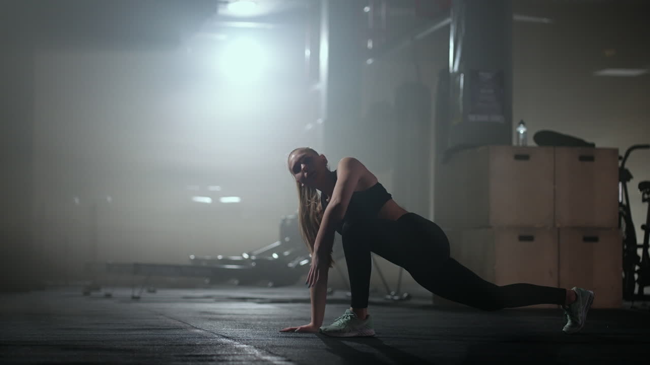 A young slender woman prepares and warms up before training. Hitching and stretching muscles after a tedious hard workout in the dark interior of the fitness room