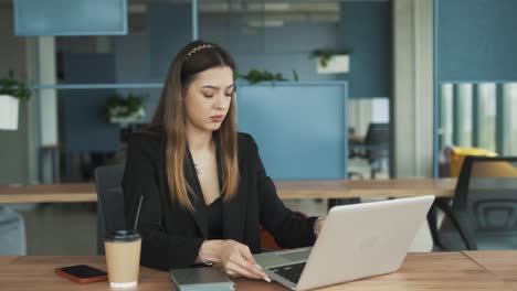 Una-Mujer-Joven-Y-Atractiva-Vestida-De-Negocios-Se-Sienta-En-Una-Elegante-Y-Moderna-Oficina-De-Coworking,-Tocando-Con-Enojo-Una-Computadora-Portátil-Que-Funciona-Mal,-Sintiéndose-Abatida