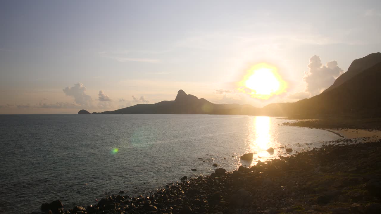 Sunset over rocky shore at Con Dao Island, Vietnam, with calm waves and a mountain backdrop