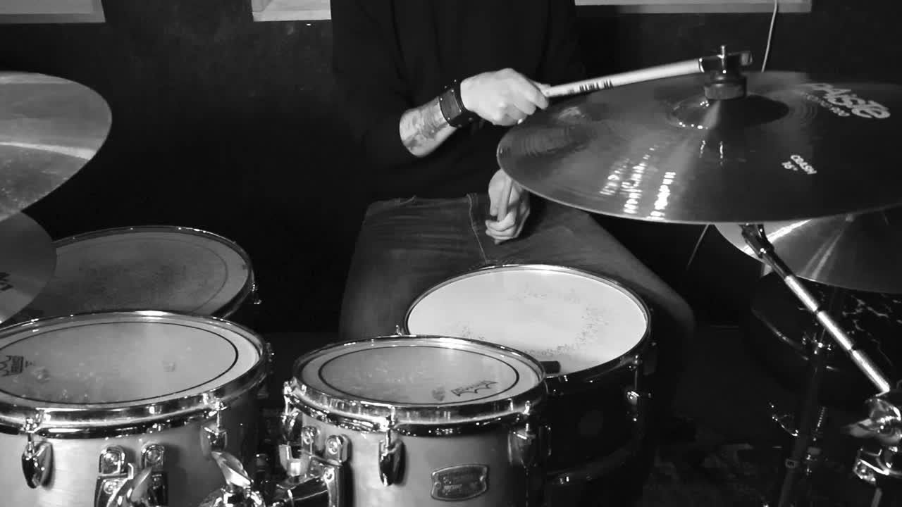 Grayscale footage of a tattooed man playing acoustic drums in slow motion in a studio room under good lights. Hitting cymbals and crashes making them move and wobble in a cinematic way.