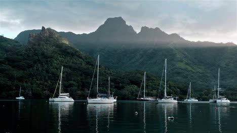 sailing yachts anchored in a beautiful and protected bay in moorea island as the sun sets behind volcanic mountain peaks