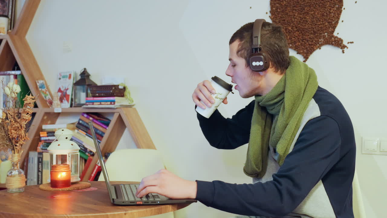Giovane uomo bianco che sorseggia caffè mentre digita sul laptop vicino a una candela e una libreria, con le cuffie e una sciarpa verde, le inquadrature mostrano uno scrittore, creatore di contenuti, editor notturno che si ferma per sorseggiare.