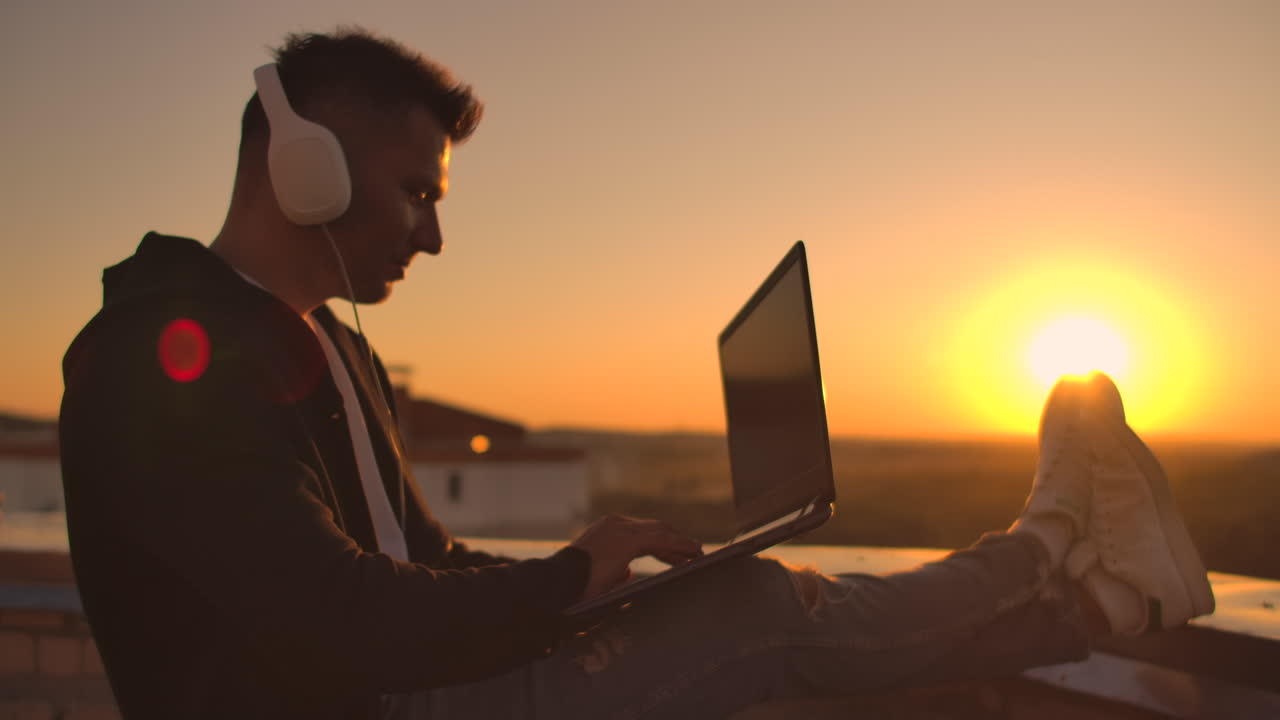 A man in headphones on the roof relaxes working remotely enjoying life despite a handsome kind of sipping beer and types on the keyboard. Trade on the stock exchange using a laptop and enjoying the beautiful view