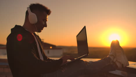 A man in headphones on the roof relaxes working remotely enjoying life despite a handsome kind of sipping beer and types on the keyboard. Trade on the stock exchange using a laptop and enjoying the beautiful view