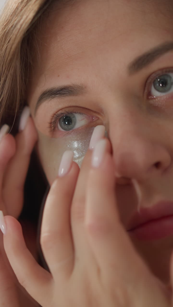 Woman strokes skin nourishing patch with finger in bathroom. Serious lady uses nourishing cosmetic product under eyes standing at mirror closeup