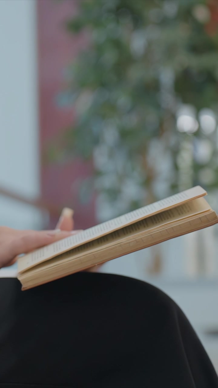 Vista laterale di una donna vestita con camicia bianca e pantaloni neri che sfoglia le pagine di un libro mentre è seduta, con uno sfondo sfocato di un centro commerciale e un'illuminazione soffusa che crea un'atmosfera di lettura tranquilla.