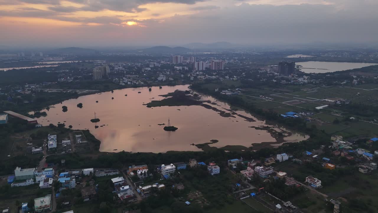A cinematic view of a developing city as day turns to night. The footage showcases a main lake, other bodies of water, and urban development in the distance under a warm, glowing sky