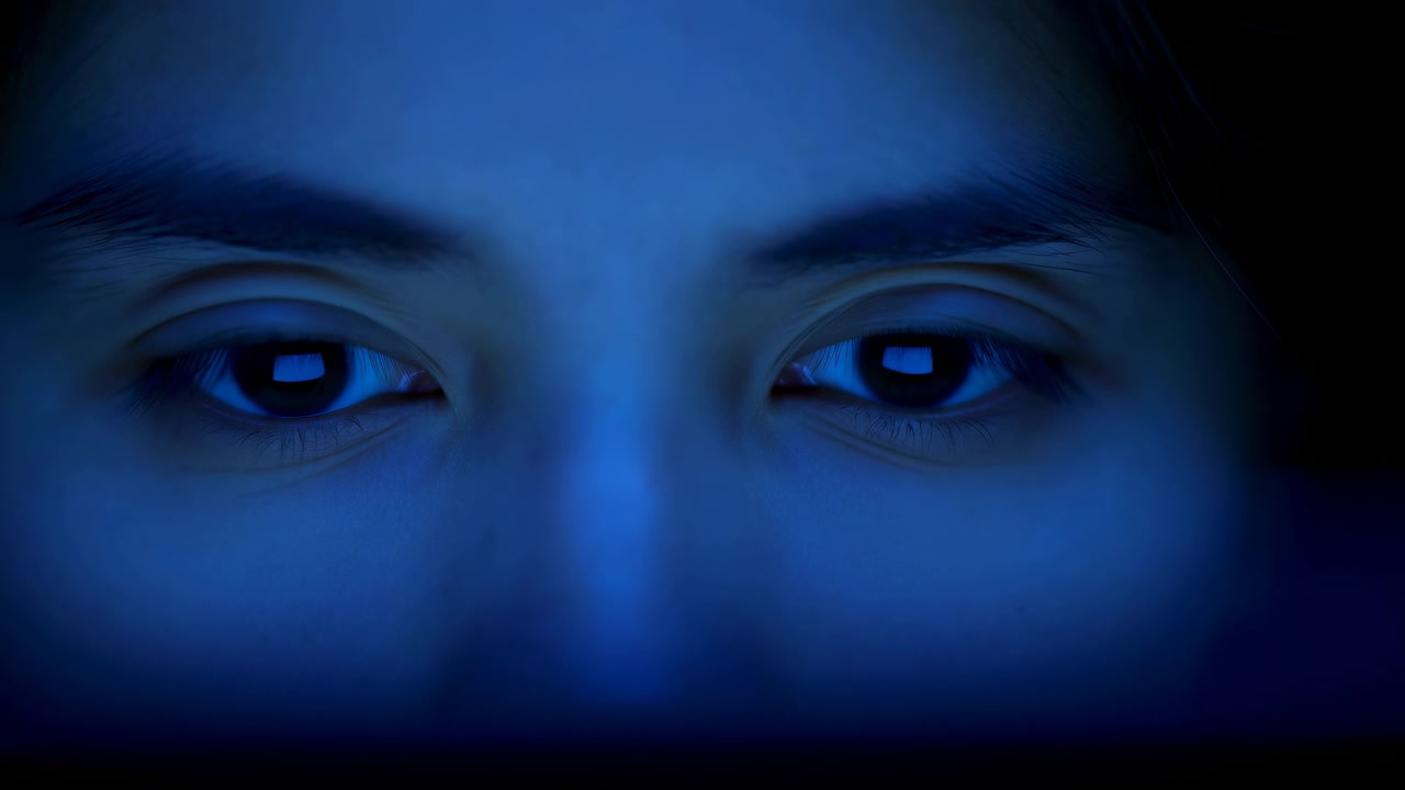 Close-up extremo mostra olhos abrindo, fechando e reabrindo em sequência, com iluminação azul fria sobre sobrancelhas e pele, arredores escuros e composição frontal estável.