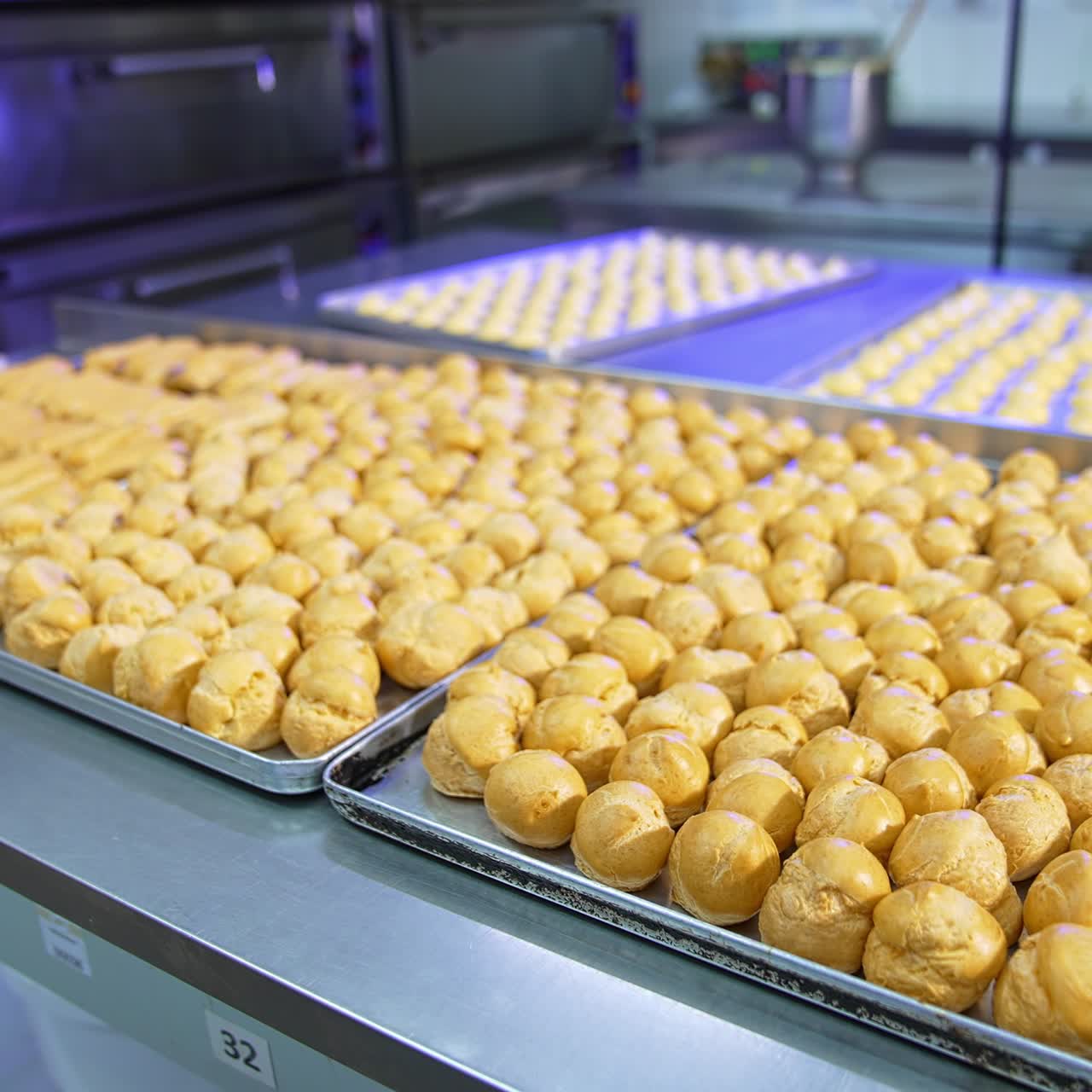 Large metal tables covered with baking sheets full of baked éclairs. Pastry prepared for stuffing with cream. Food factory production