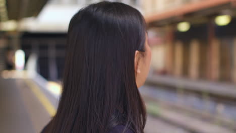 close up rear view of young woman waiting on railway station platform for train looking at mobile phone