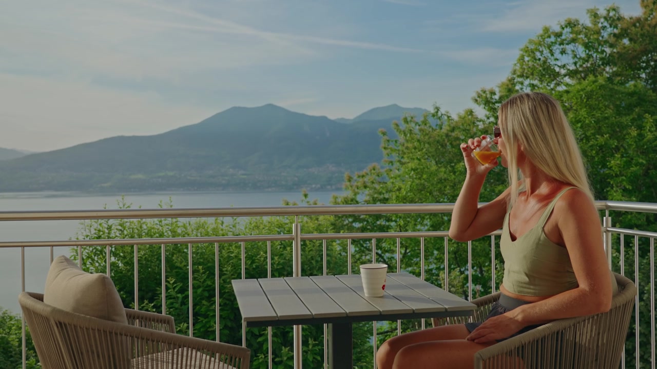 Una donna si rilassa su un balcone di un resort di lusso in riva al lago con vista sul Lago Maggiore, sorseggiando succo d'arancia fresco mentre si gode una vista mozzafiato sulle montagne in una splendida giornata estiva italiana.