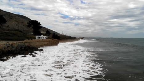 Aerial-drone-shot-on-a-cloudy-day-over-the-Pacific-Ocean-waves-next-to-RV-campers-and-the-cliffs-along-the-101-freeway-in-Ventura,-California