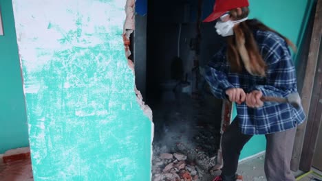 joven mujer rubia con mascarilla demoliendo muro de hormigón usando mazo - plano medio