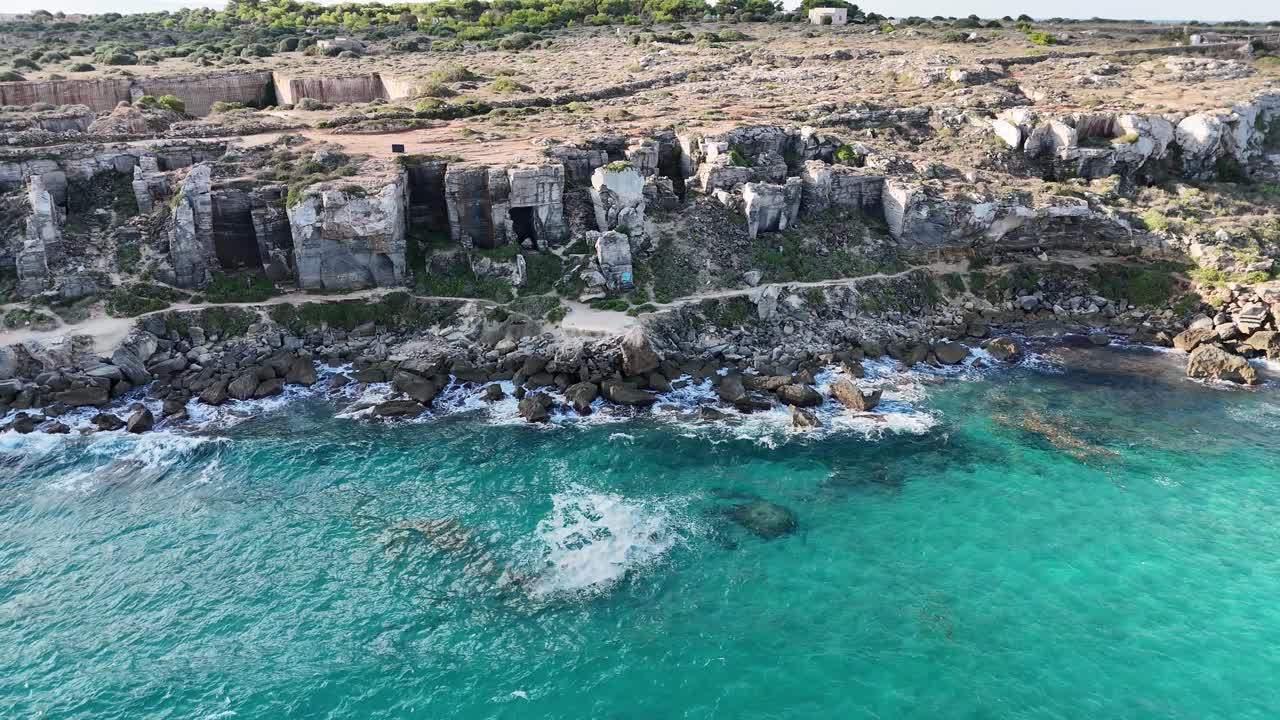Fly-right aerial view of Cala Rossa’s turquoise water, rocky coastline, and tuff quarry cliffs in Favignana, Sicily. Rough waves crash against the shore during a mid-morning pass