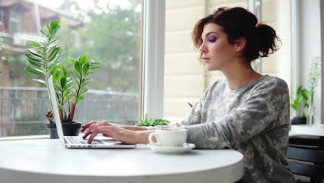 Mujer-Hermosa-Joven-Sentada-Cerca-De-La-Ventana-En-La-Cafetería-Y-Trabajando-En-Una-Computadora-Portátil.-Chica-Navegando-Por-Internet-Y-Escribiendo