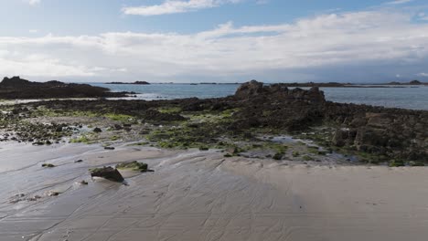 Low-drone-flight-over-beach-rockpools-and-rocks-at-low-tide-out-to-sea-from-Belle-Greve-Bay-Guernsey-on-bright-calm-day