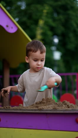 happy caucasian child plays with sand outdoors. lovely toddler uses a shovel to push the sand. vertical video.