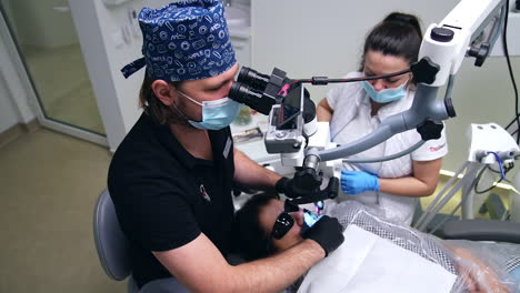 male doctor in blue cap and mask looking at binoculars of endodontic microscope. dentist working in the mouth of patient wearing black glasses. view from top.