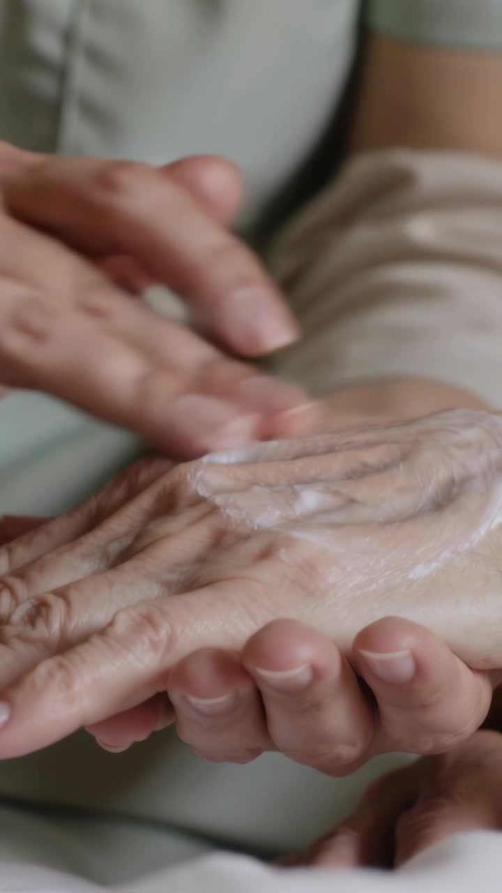 Vertikales Video: Eine Pflegekraft in hellem Oberteil trägt Creme auf und reibt die Hand eines Seniors auf dem Bett zur Hautpflege.