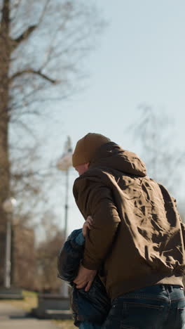 padre levantando a su hijo ambos están vestidos con vaqueros y con el niño con una chaqueta hinchada y el hombre con un gorro, árboles y un poste eléctrico en el fondo