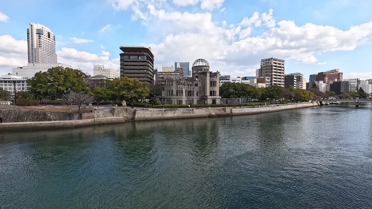 Wide view of the Hiroshima Peace Memorial Dome along the Motoyasu River in Hiroshima, Japan, with urban skyline and trees lining the riverbank under a blue sky.