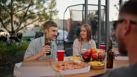 Ein-Glücklicher-Blonder-Typ-Mit-Einer-Flasche-Bier-In-Der-Hand-Kommuniziert-Mit-Seinen-Freunden-Und-Hat-Spaß-Beim-Mittagessen-Im-Hof-Eines-Landhauses-Am-Tisch