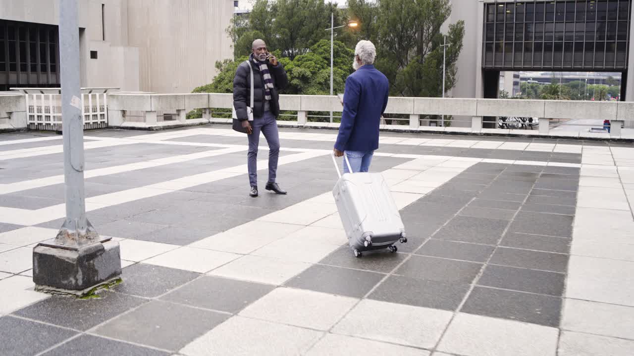 African American men, one waving phone cue to guide other towing suitcase across plaza