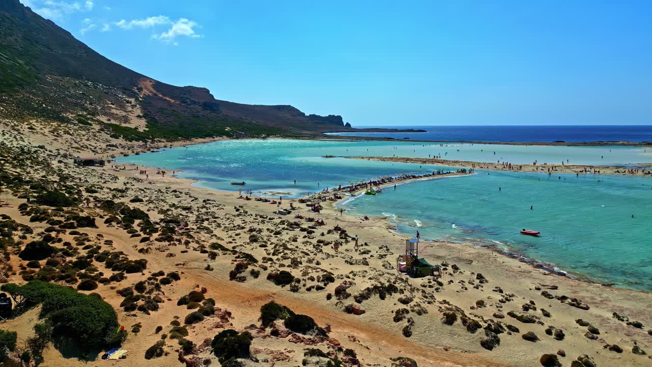 Ripresa aerea con drone che avanza sopra la spiaggia di Balos a Creta, Grecia. Una vasta laguna turchese incontra rive sabbiose dorate, con bagnanti, scogliere rocciose e il blu profondo del Mediterraneo in lontananza.