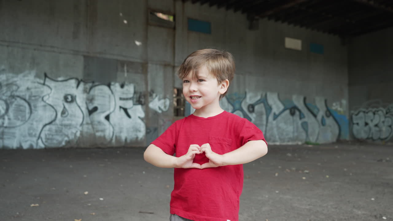 A young little boy is having fun and making heart symbols with hands in front of camera in a red T-shirt. Camera rotating around a young boy standing in front of a graffiti wall at a abandoned place.
