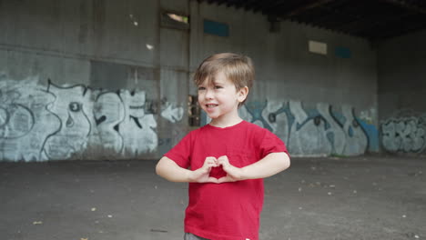 a young little boy is having fun and making heart symbols with hands in front of camera in a red t-shirt. camera rotating around a young boy standing in front of a graffiti wall at a abandoned place.