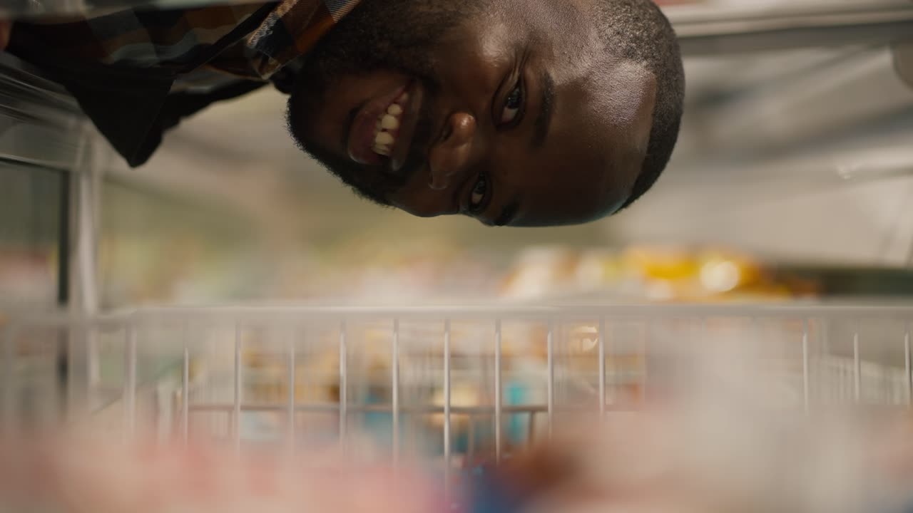 Close-up shot of a Black-skinned man in a plaid shirt looks into the supermarket refrigerator, looks into the camera and takes an ice cream