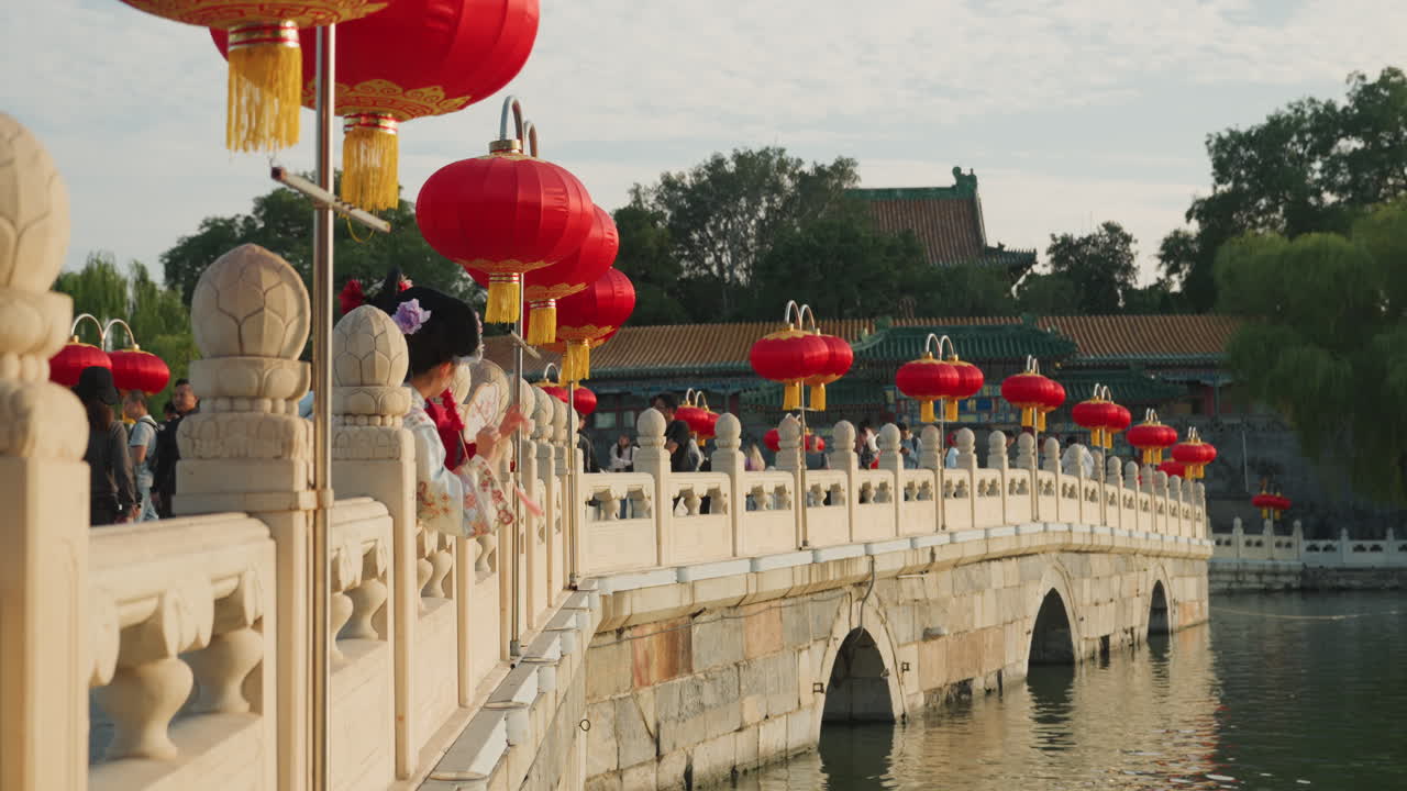 Đèn lồng trên cầu đá lúc hoàng hôn với đám đông tản bộ dọc theo lan can trang trí công phu, đèn lồng đỏ treo trên cột, mặt nước tĩnh lặng phản chiếu vòm cong, mái chùa truyền thống và cây cối