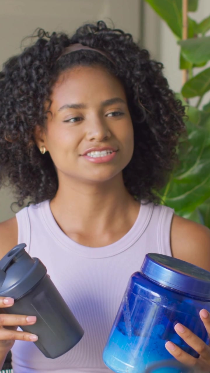 Vídeo vertical: Mulher de regata falando, mostrando um pote azul e coqueteleiras pretas e verdes para uma demonstração em casa.